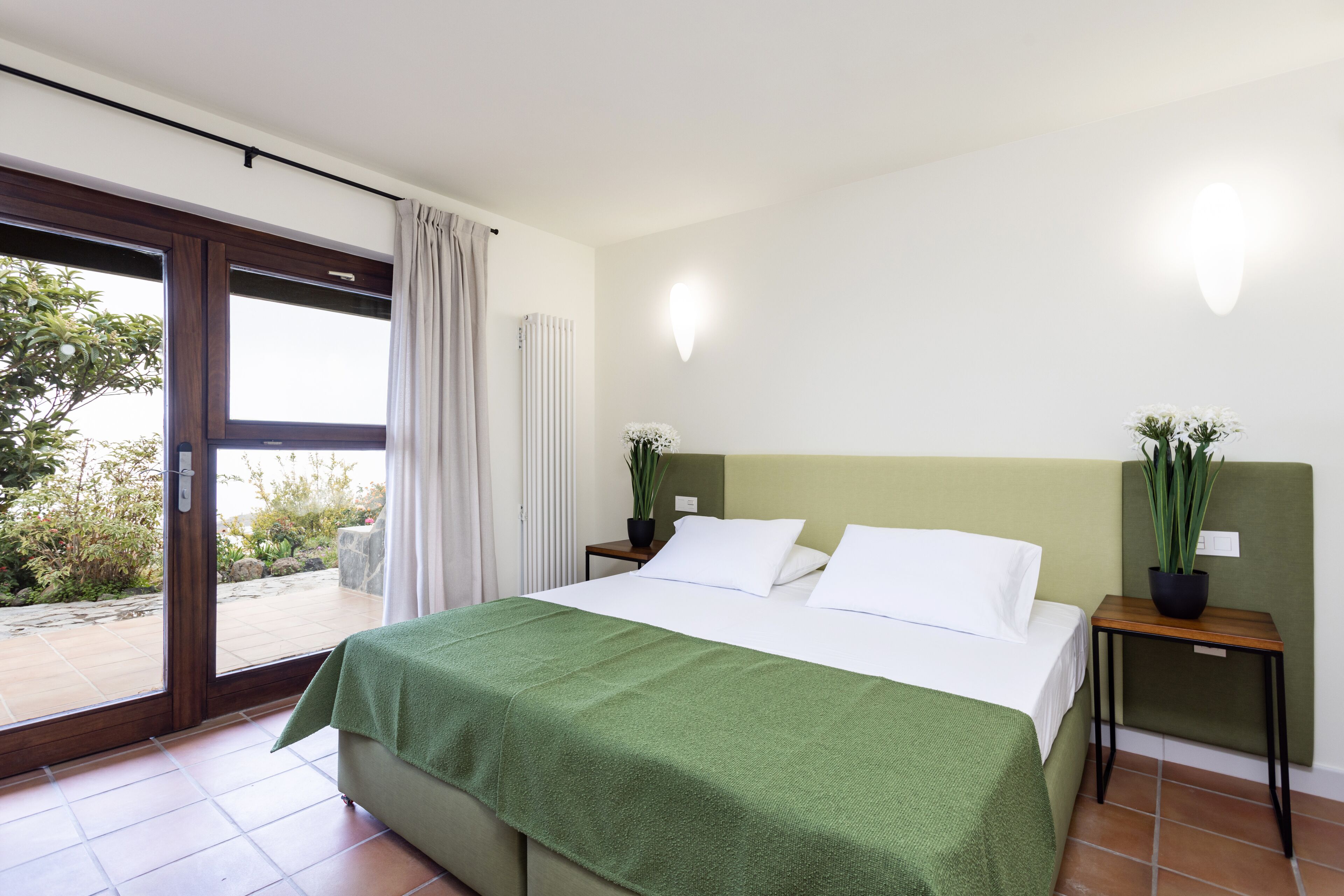 Habitación de hotel con cama, manta verde, dos plantas en macetas, una mesita de noche y una puerta corrediza de vidrio que da a un espacio al aire libre.