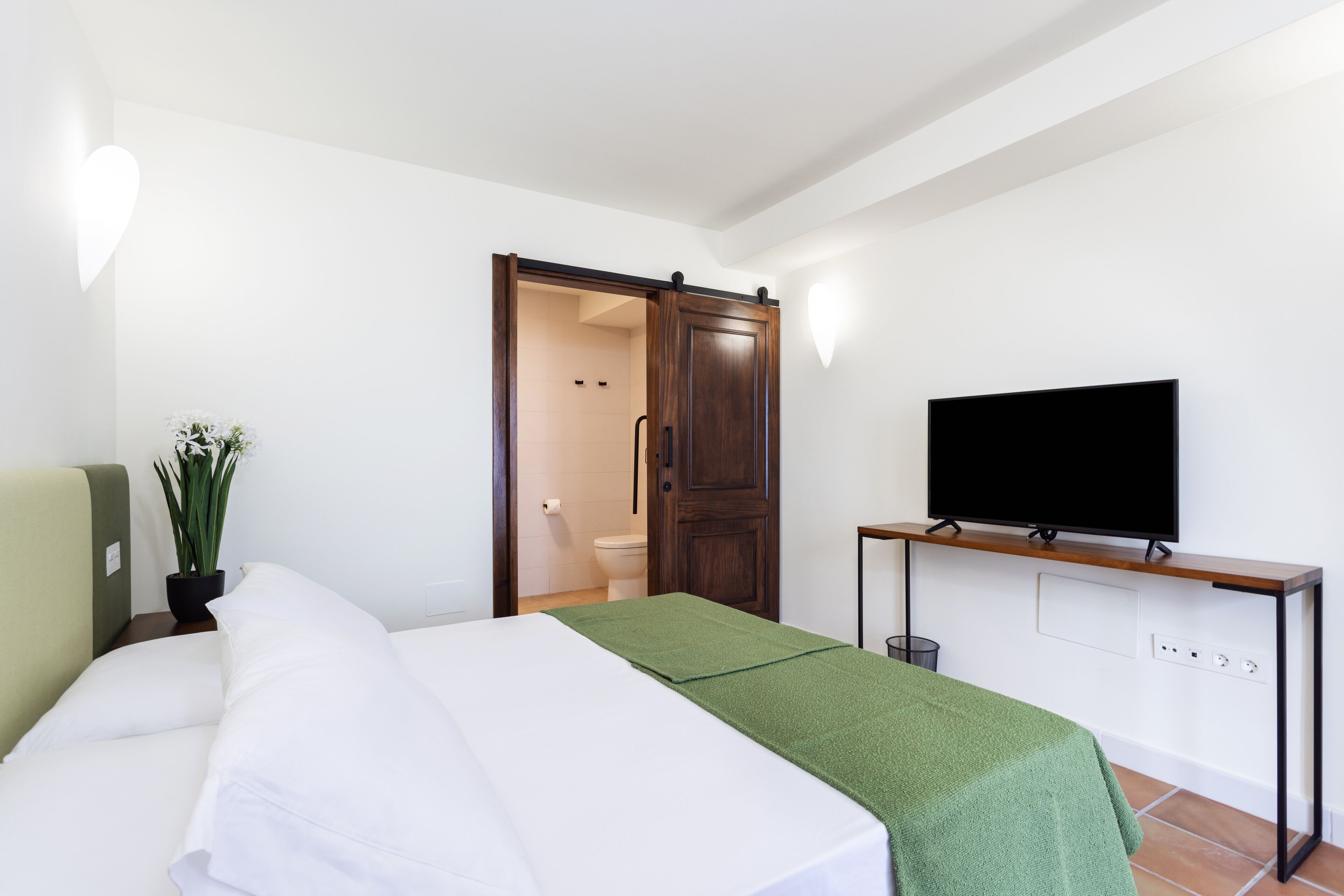A hotel room with a bed, a TV on a stand, a green towel, and a wooden wardrobe.