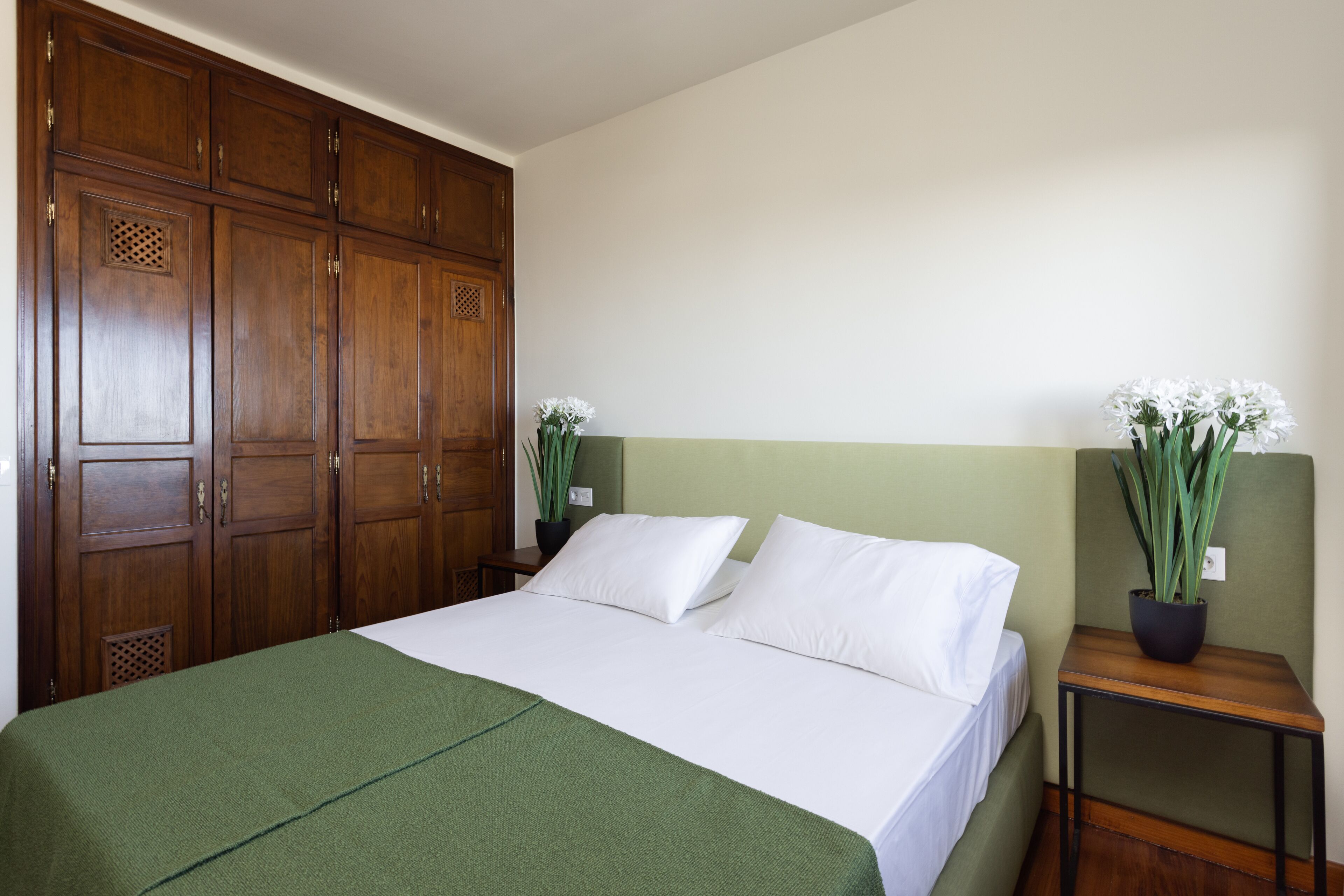 Una cama bien hecha con una manta verde, un armario de madera y una mesita con una planta en maceta.