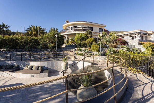 A modern building with a curved balcony, a pool area with a seating area, and a well-maintained garden with various plants. - Hotel Alta Montaña (Vilaflor)