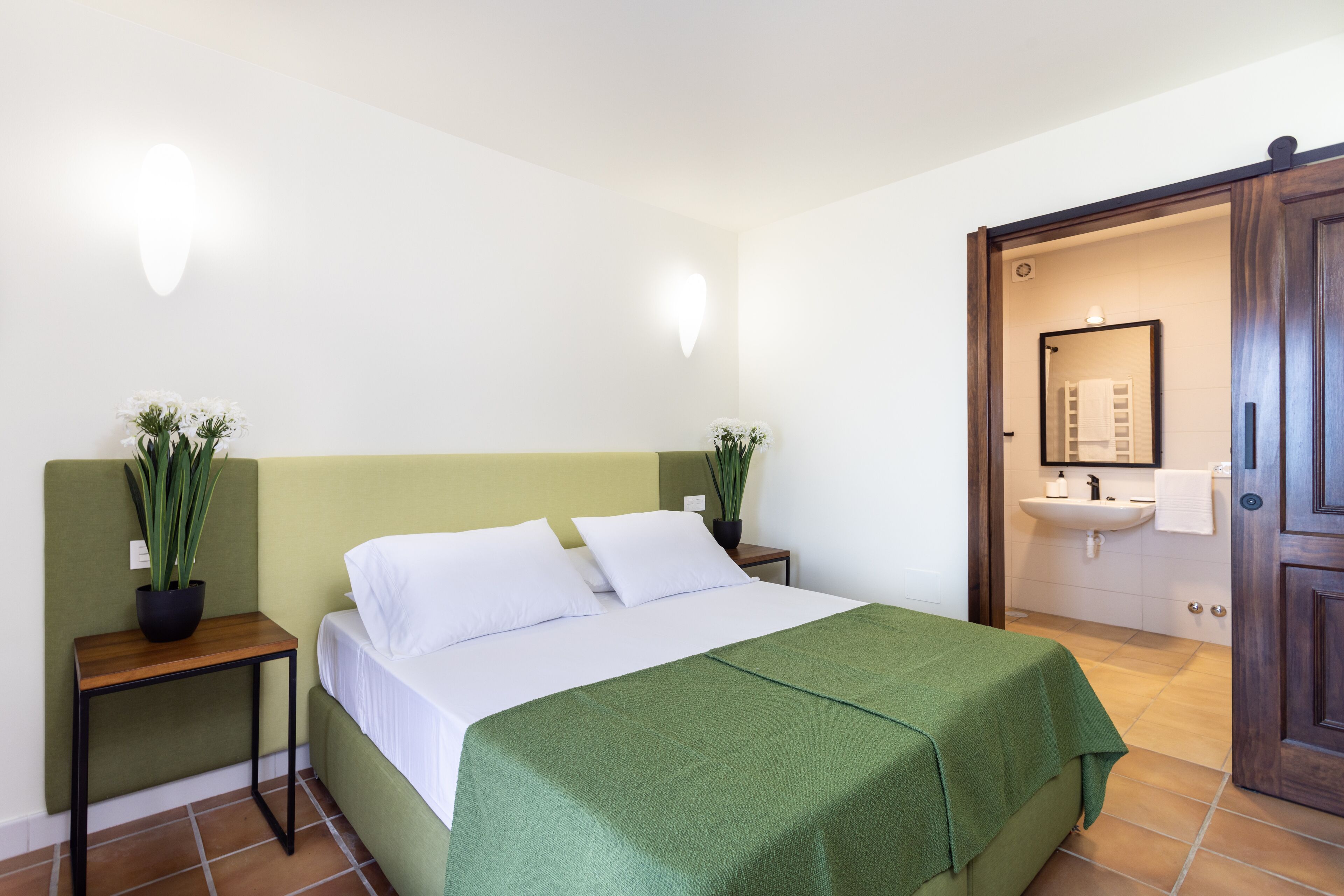 A neatly arranged bedroom with a bed, bedside tables, a green bedspread, and potted plants.