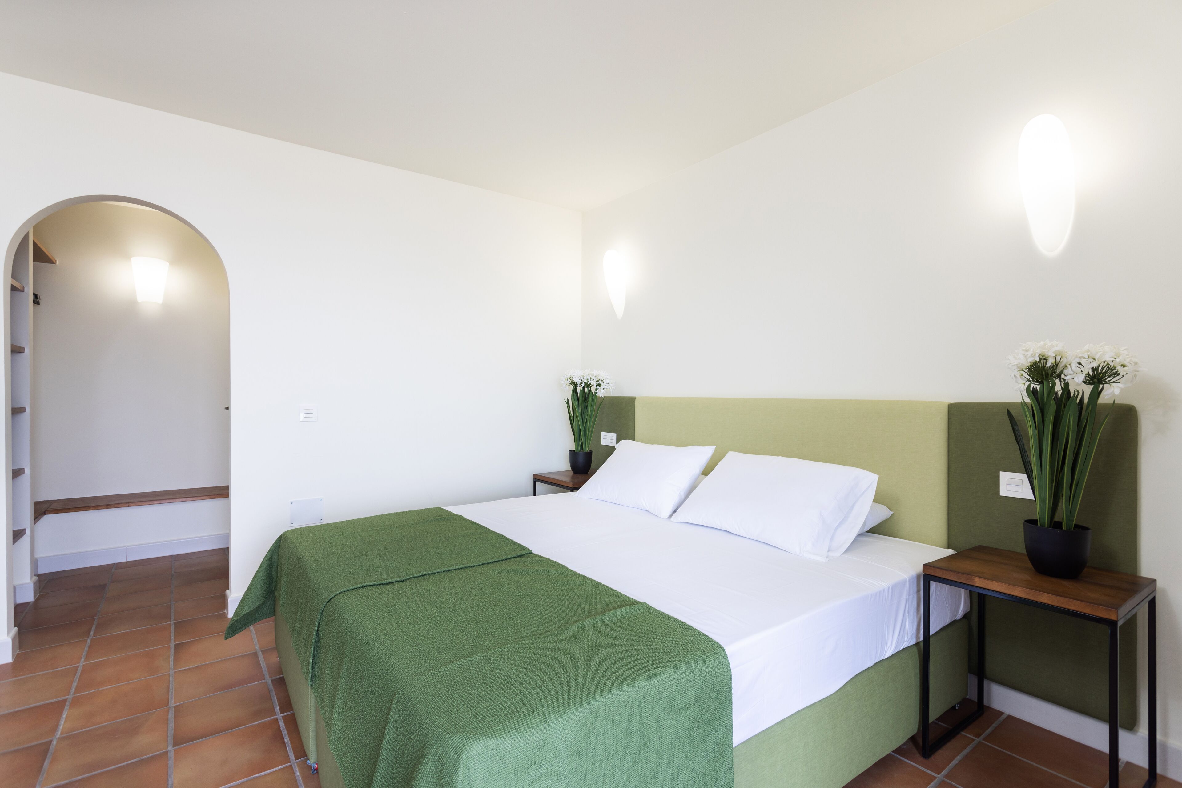 A neatly made bed with a green bedspread, a bedside table with a plant, and a wall-mounted light fixture.