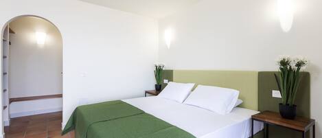 A neatly made bed with a green bedspread, a bedside table with a plant, and a wall-mounted light fixture.