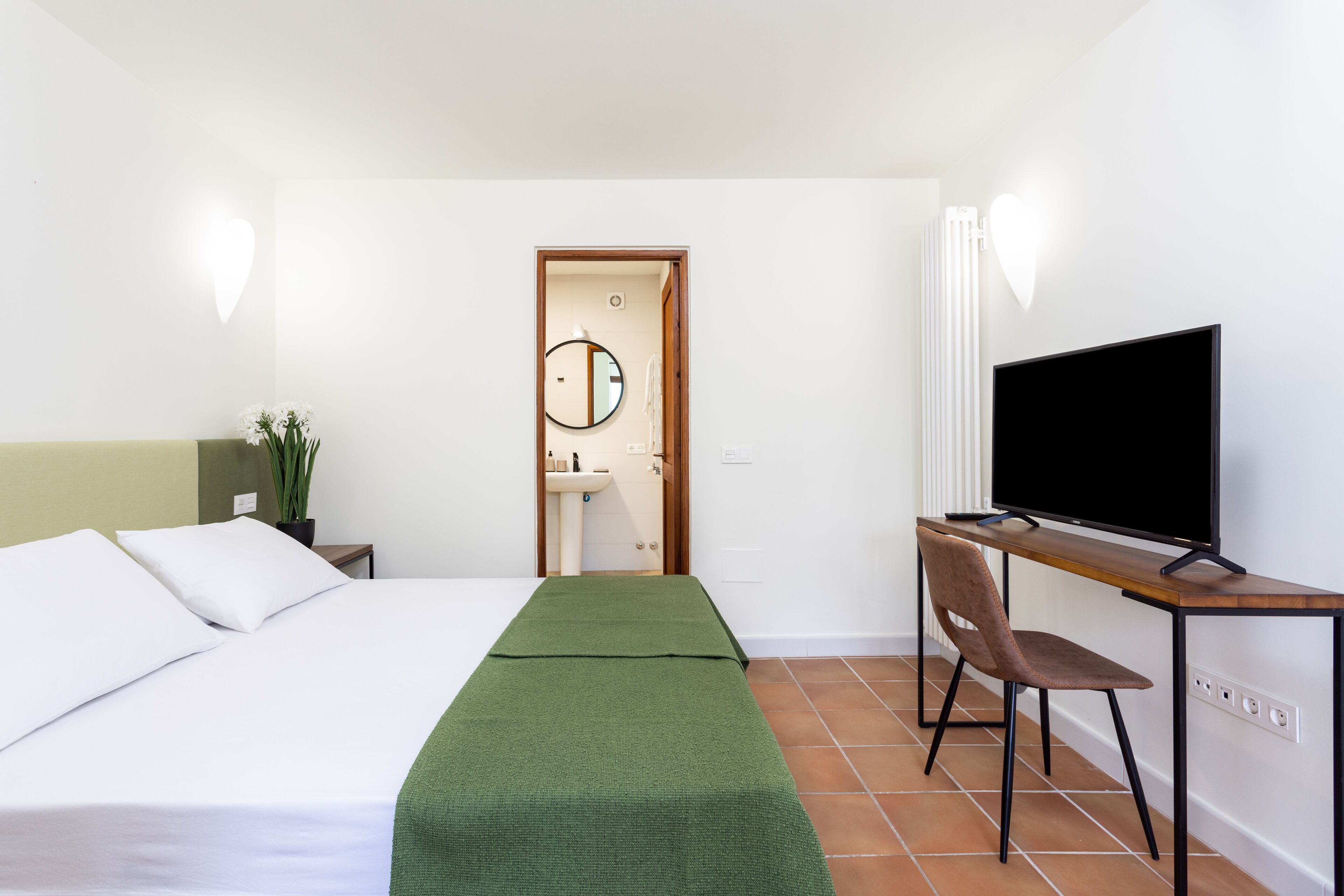 Ein Hotelzimmer mit Bett, Schreibtisch mit Stuhl, Fernseher, Spiegel und einer Pflanze.