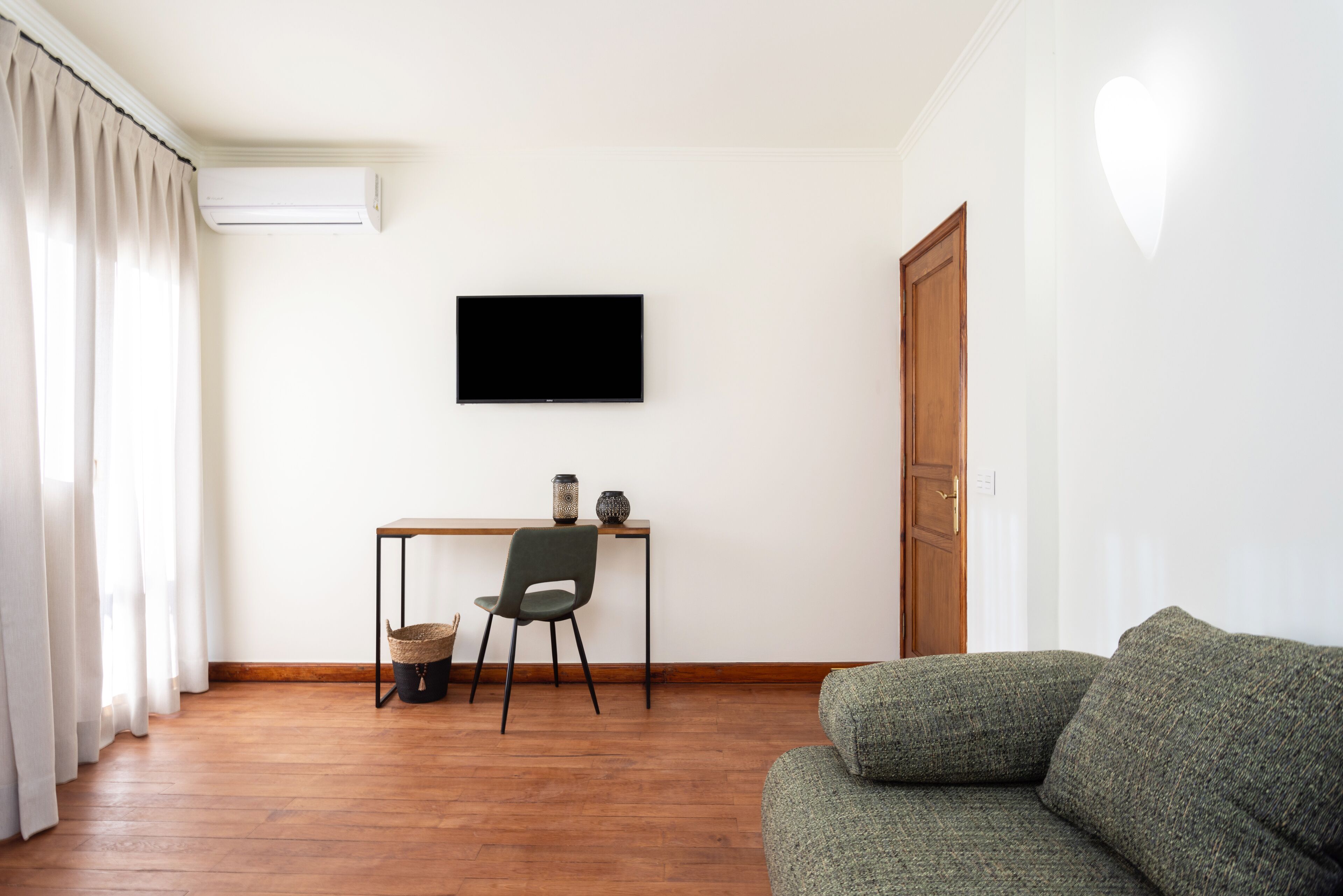 Una sala de estar con piso de madera, un sofá verde, un sillón negro, una mesita con objetos decorativos y un televisor montado en la pared.