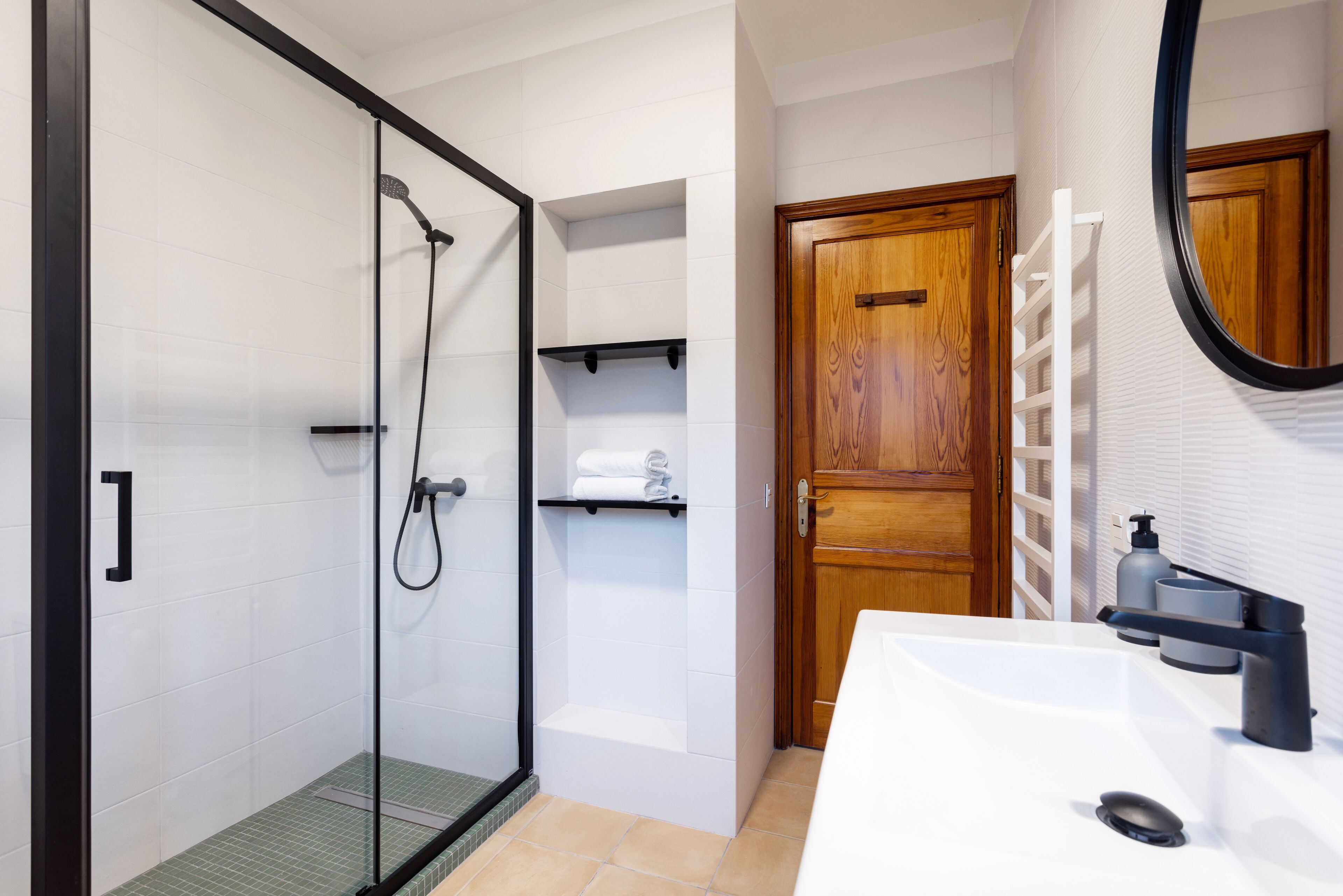 Une salle de bain moderne avec une douche à parois de verre, un lavabo blanc doté d’un robinet noir et une porte en bois.