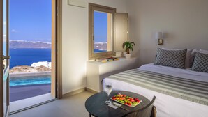 Habitación de hotel con una cama, una mesita de noche con una bandeja de fruta, un vaso de agua y vistas al mar.