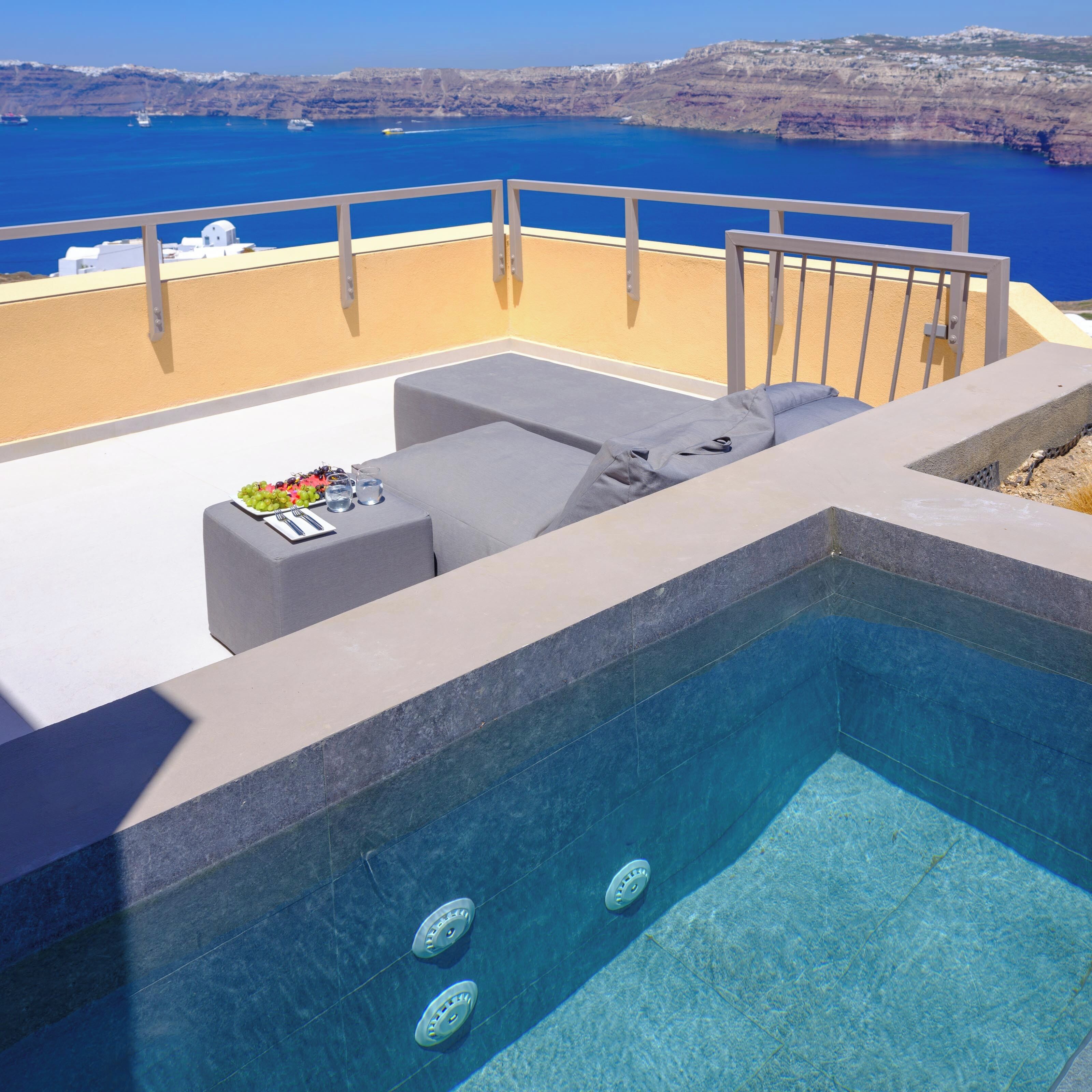 A rooftop pool with clear blue water, a seating area with cushions, and a view of the sea and cliffs.