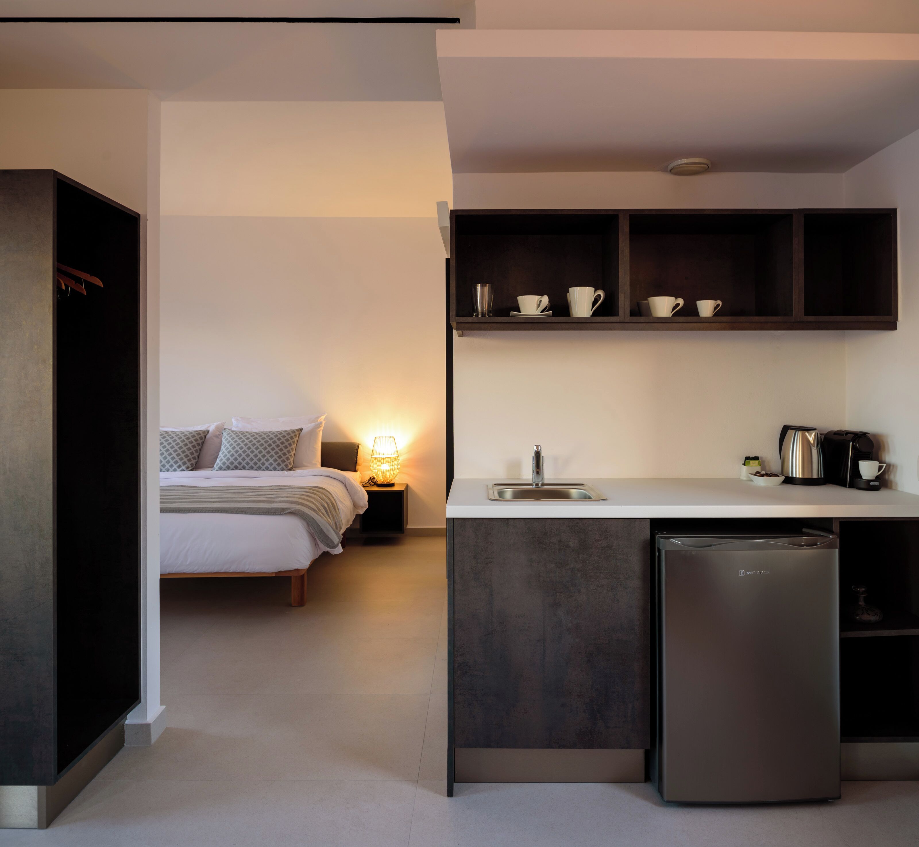 A compact hotel room with a kitchenette, a bed with patterned pillows, a bedside lamp, and a small cabinet above the sink.