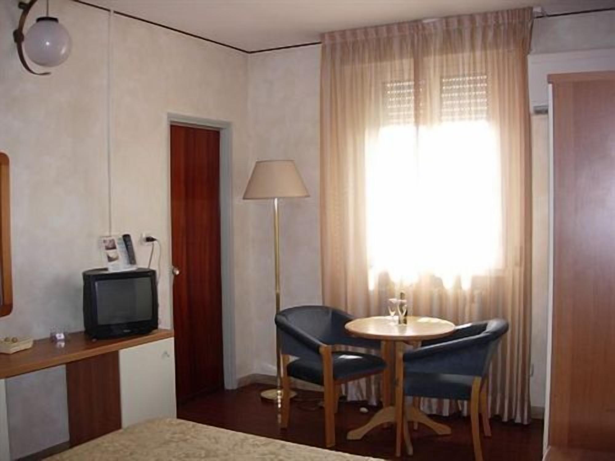 Habitación de hotel con cama, escritorio con televisor, silla, mesa, lámpara y ventana con cortinas.