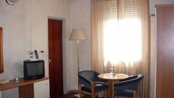 Habitación de hotel con cama, escritorio con televisor, silla, mesa, lámpara y ventana con cortinas.
