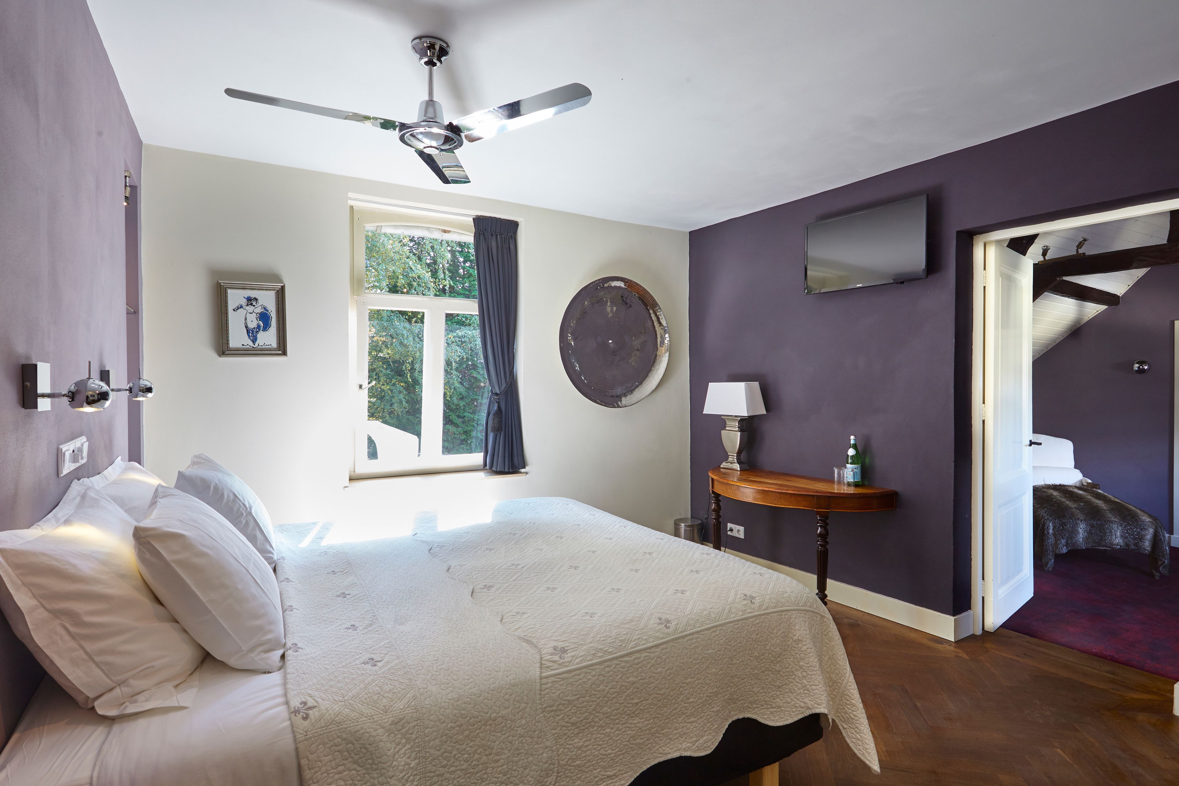 A bedroom with a bed, a ceiling fan, a window with curtains, a round mirror, a small table with a lamp, and a TV mounted on the wall.