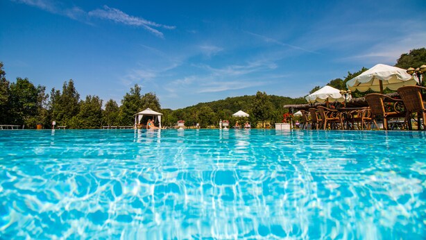 Ein klares, blaues Schwimmbecken mit Liegestühlen und Sonnenschirmen, umgeben von Bäumen und einer Hügelkulisse im Hintergrund.