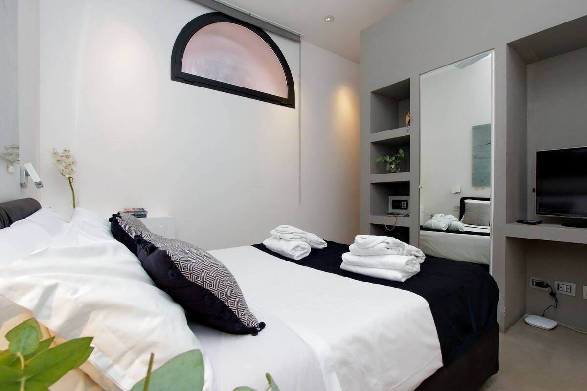 A modern hotel room with a bed, a TV, a mirror, and a shelf with toiletries.