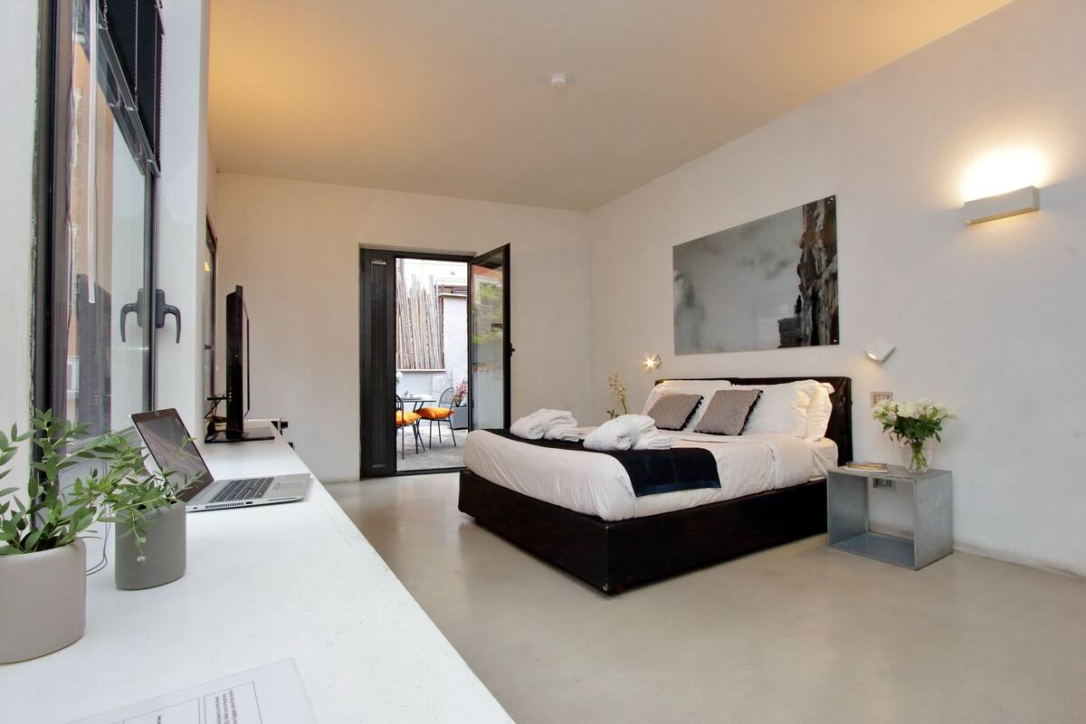 A modern bedroom with a large bed, a desk with a laptop, and a potted plant.