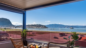 A balcony with wicker chairs, a table with breakfast items, and a view of a coastal landscape with mountains and a bridge. - Harmony Hotel Nafplio (Nafplio)