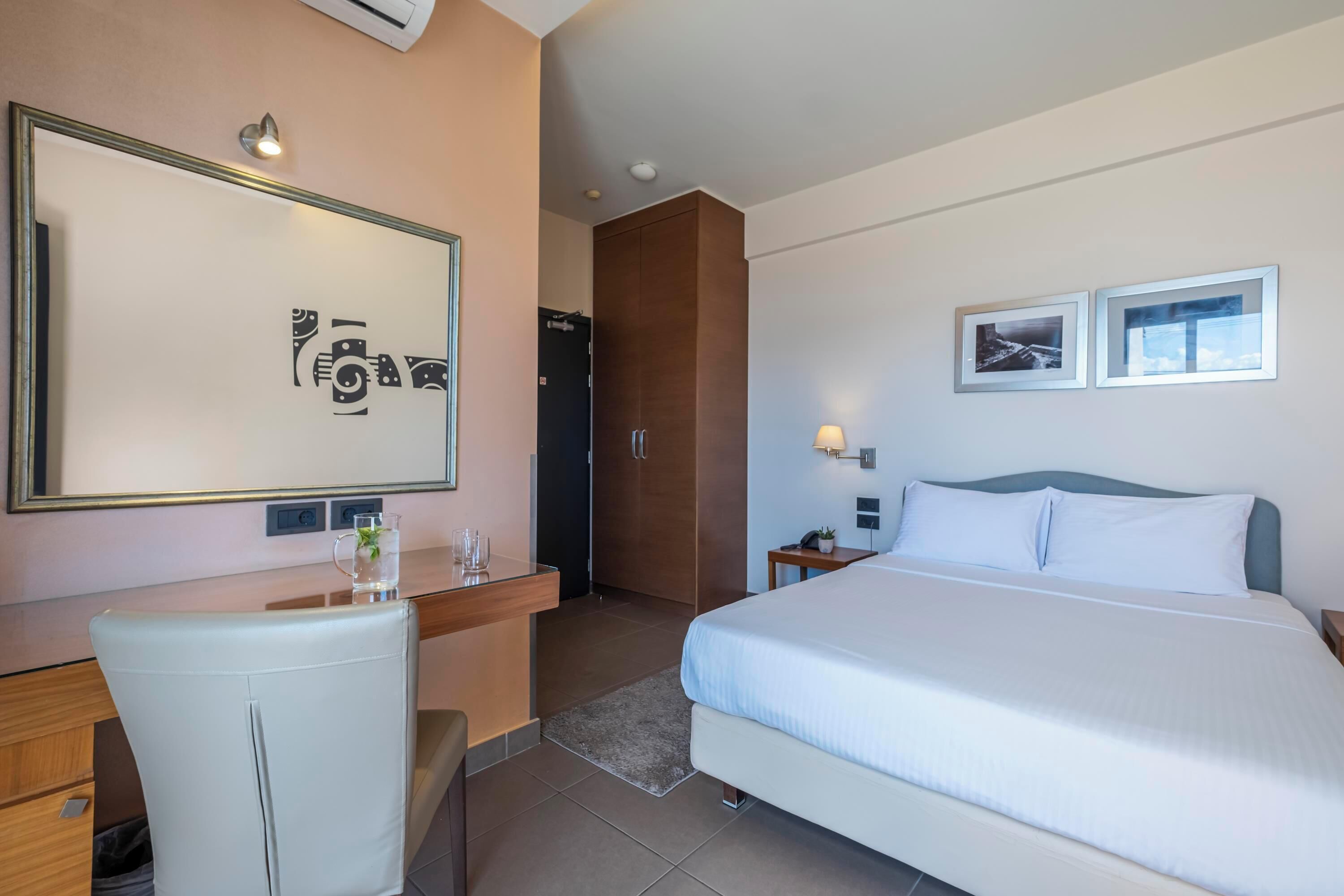 A hotel room with a bed, a desk, a chair, a mirror, and two framed pictures on the wall.