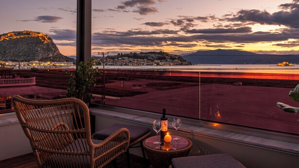 A balcony with wicker furniture, a bottle of wine, and a glass, overlooking a waterfront with a distant mountain and cityscape. - Harmony Hotel Nafplio (Nafplio)