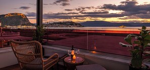 A balcony with wicker furniture, a bottle of wine, and a glass, overlooking a waterfront with a distant mountain and cityscape. - Harmony Hotel Nafplio (Nafplio)
