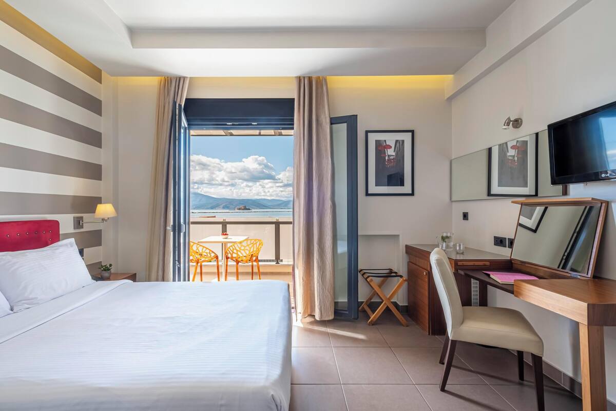 Habitación de hotel con cama, escritorio, silla, televisor y balcón con vista al mar.