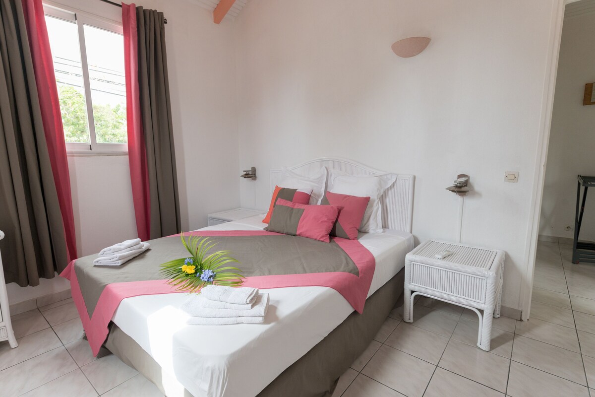 Ein ordentlich bezogenes Bett mit einer rosa und grauen Tagesdecke, einem weißen Kopfteil und einem kleinen weißen Nachttisch daneben.