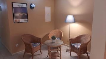 A room with two wicker chairs, a small round table, a floor lamp, a wall-mounted light, and a framed picture on the wall.