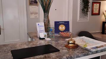 Un mostrador de recepción de hotel con una zona de registro, un jarrón con plantas altas, un premio enmarcado y un certificado en la pared.