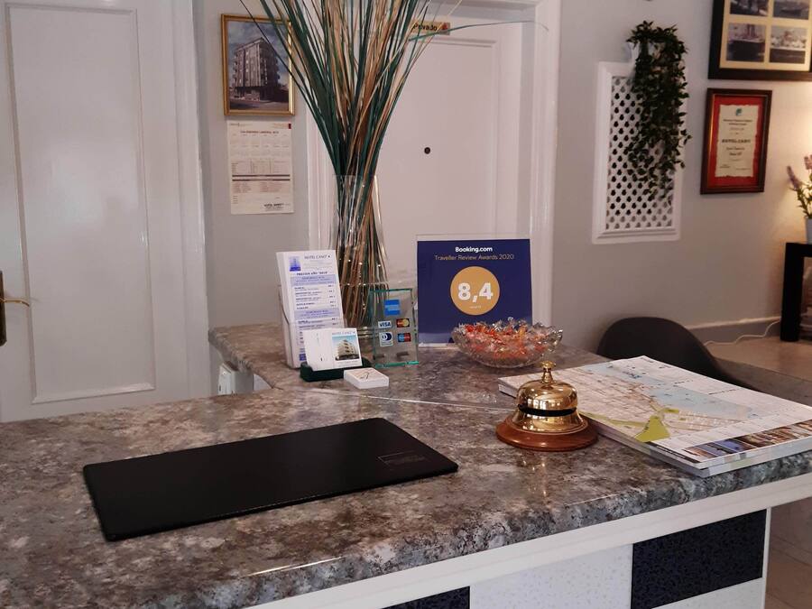 Un mostrador de recepción de hotel con una zona de registro, un jarrón con plantas altas, un premio enmarcado y un certificado en la pared.