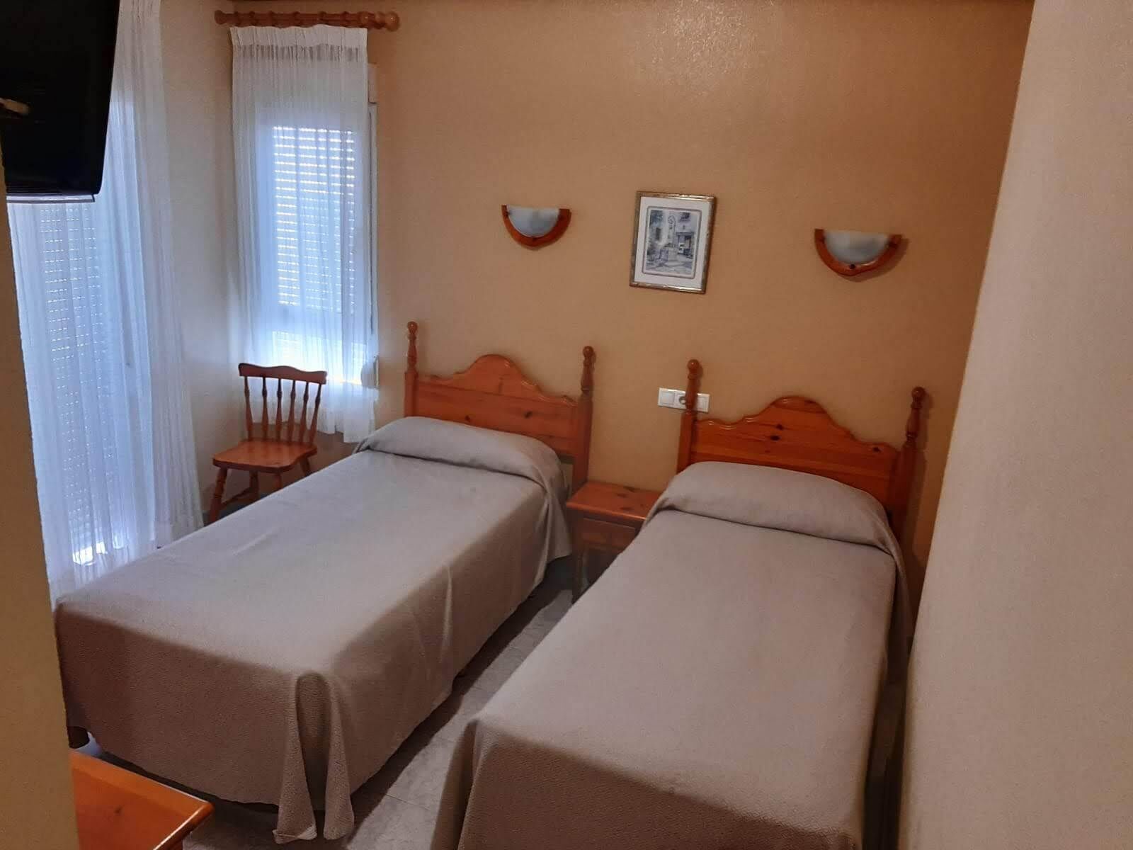 A room with two single beds, a chair, a small table, and a window with curtains.