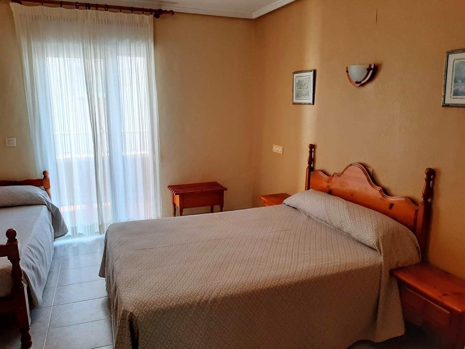 Un dormitorio con una cama grande, una cama más pequeña, una mesita de noche y una ventana con cortinas translúcidas.