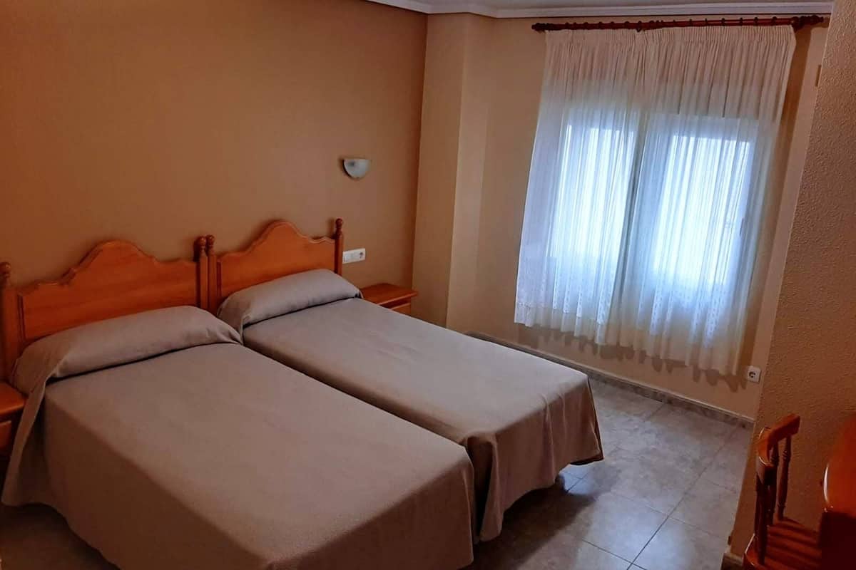 Una habitación con dos camas, cabecero de madera, una mesita de noche, un reloj de pared y una ventana con cortinas translúcidas.