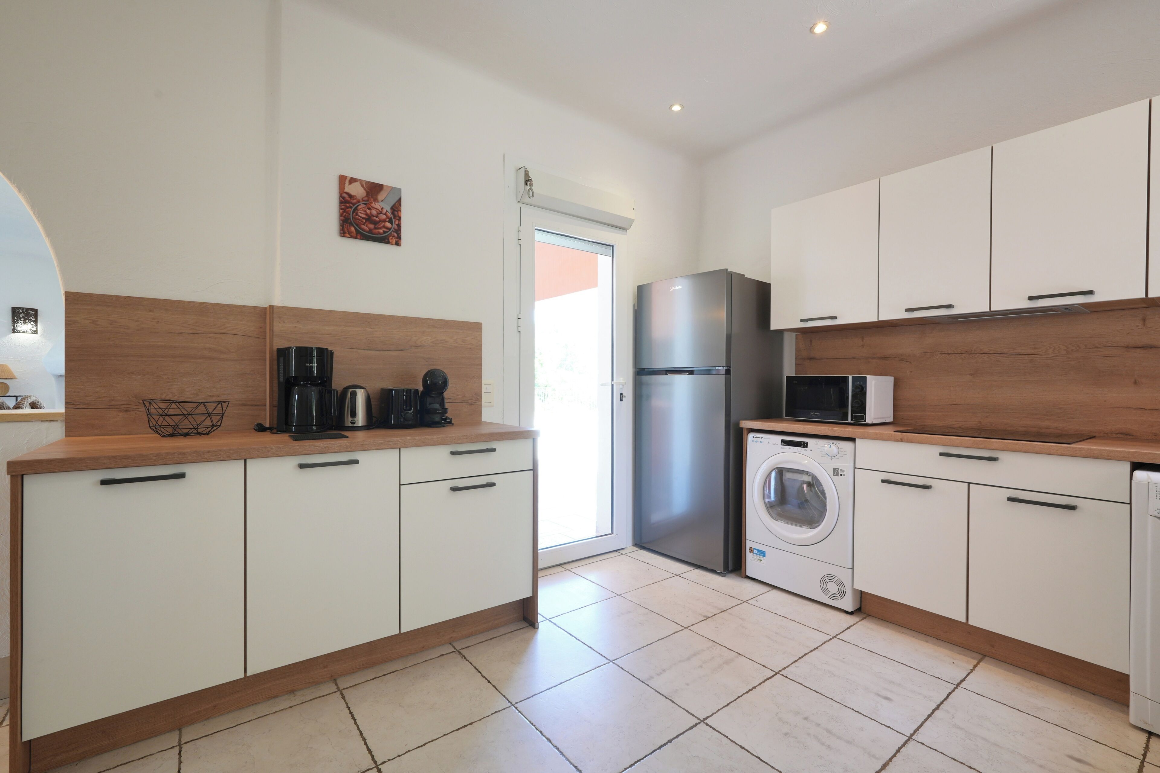 A modern kitchen with white cabinets, a stainless steel refrigerator, a microwave, a coffee maker, and a washing machine.