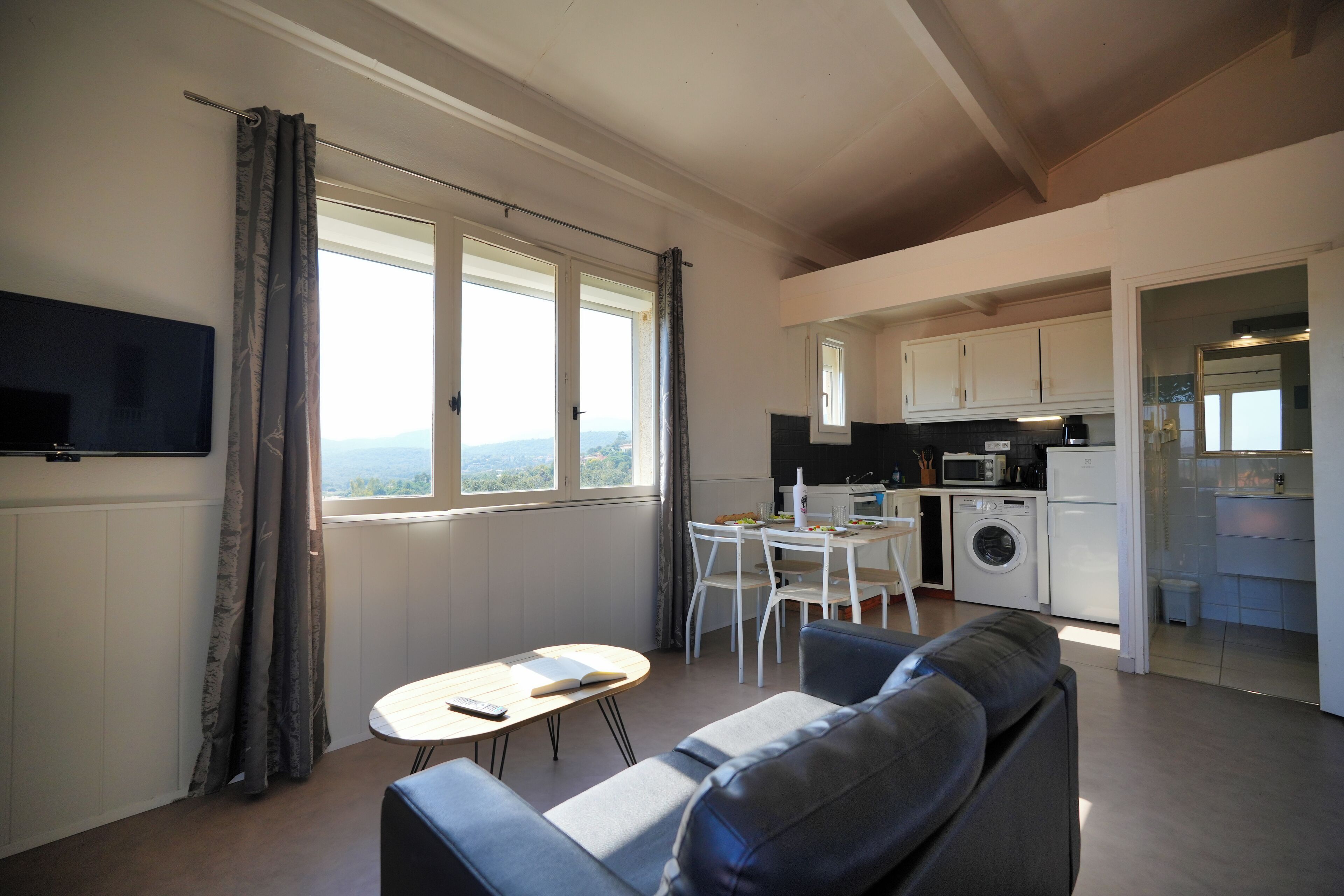 A living room with a sofa, a small table, a TV mounted on the wall, and a view of the kitchen and outside through large windows.