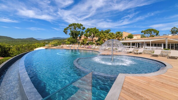 A large, curved swimming pool with a wooden deck and lounge chairs, surrounded by trees and buildings. - Hotel Residence U Paesolu (Zonza)