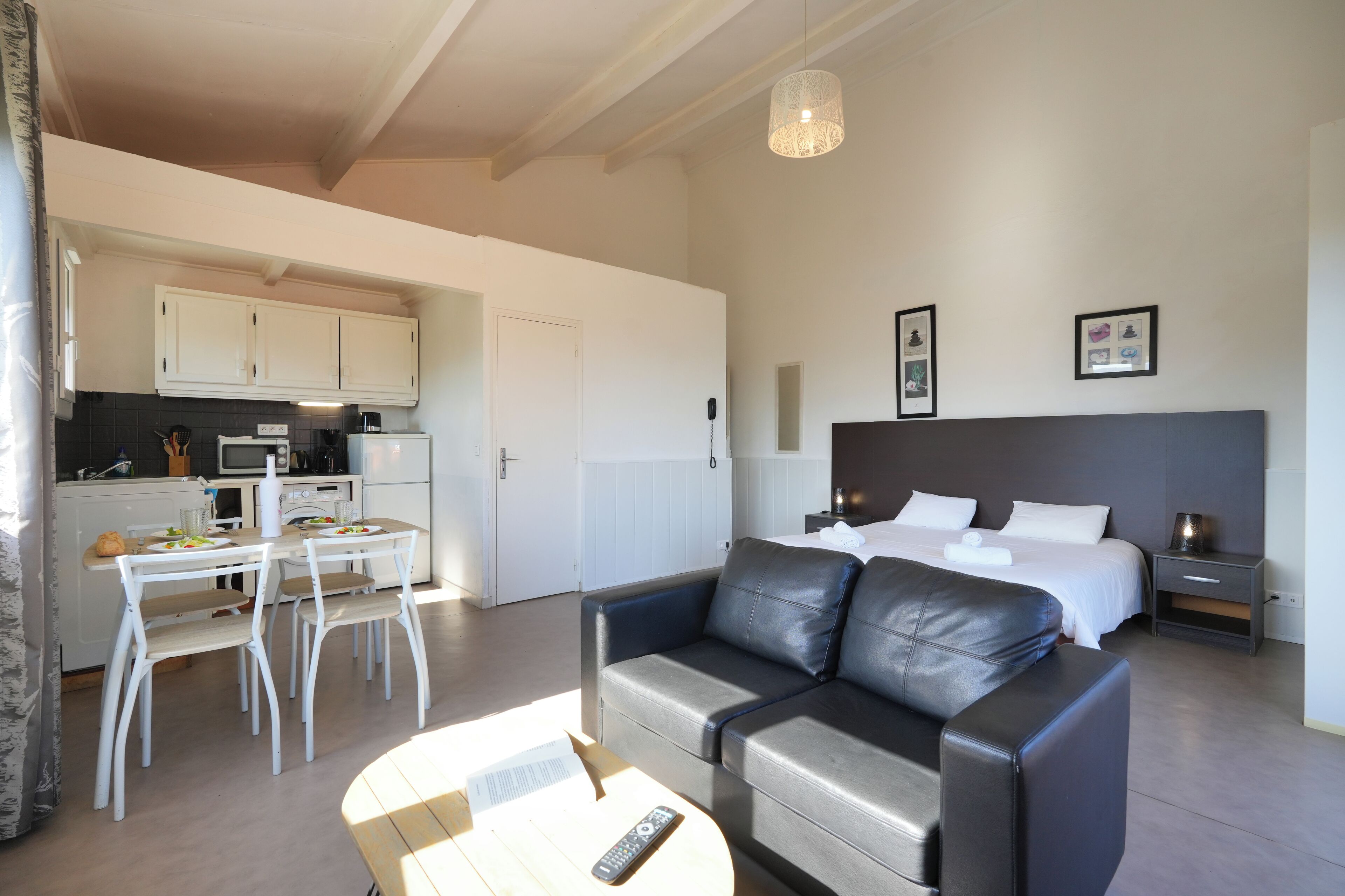 Habitación de hotel moderna con cama, sofá, mesa de comedor con sillas, zona de cocina y lámpara colgante.