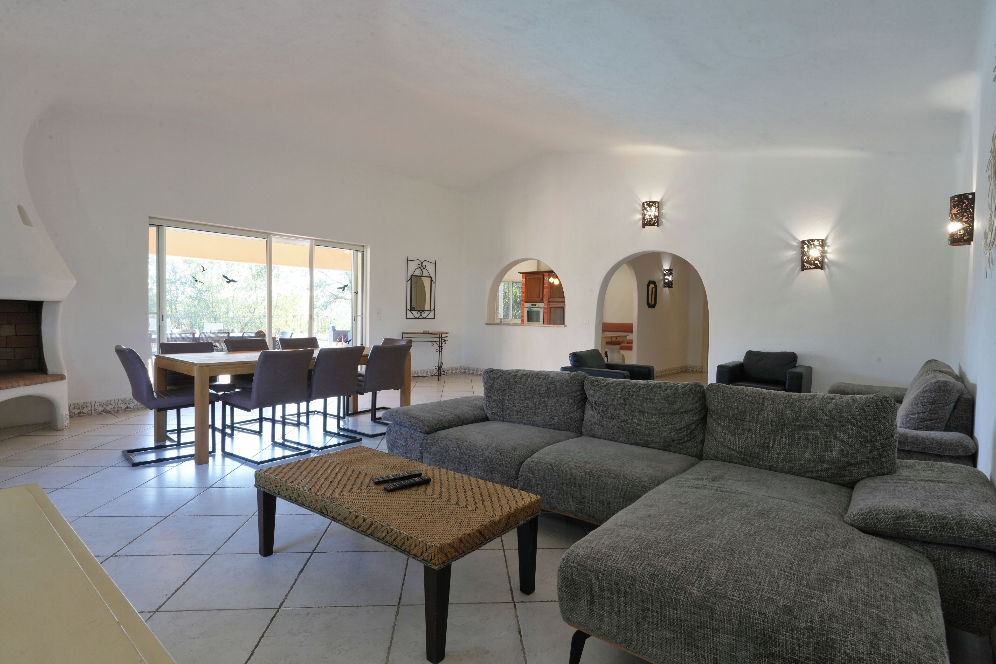 A spacious living area with a large gray sectional sofa, a small woven coffee table, and a dining table with chairs. There are wall-mounted sconces providing ambient lighting.