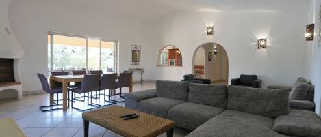 A spacious living area with a large gray sectional sofa, a small woven coffee table, and a dining table with chairs. There are wall-mounted sconces providing ambient lighting.