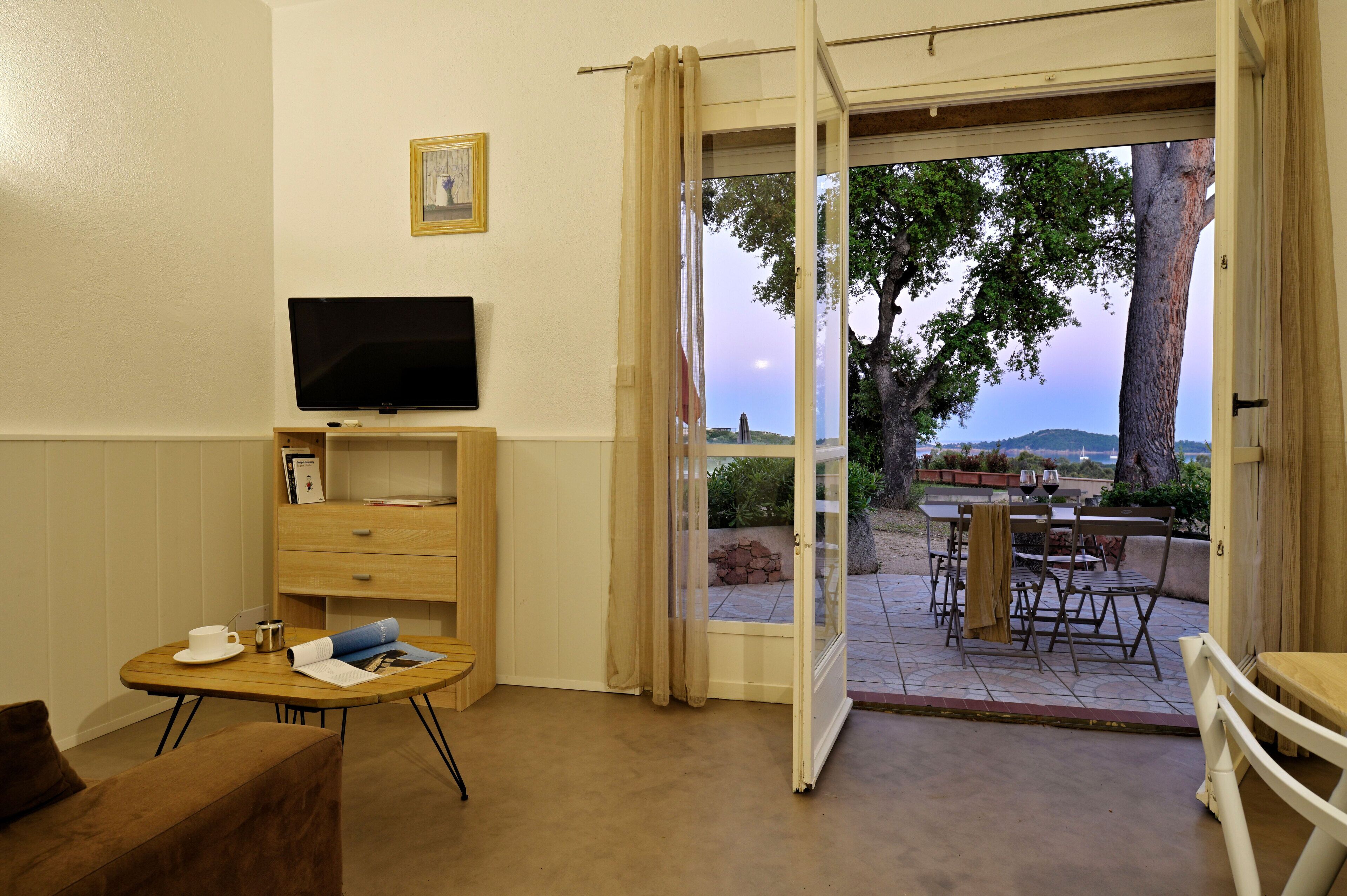 A room with a television on a wooden stand, a small table with a cup and papers, and an open door leading to an outdoor patio with a table and chairs.