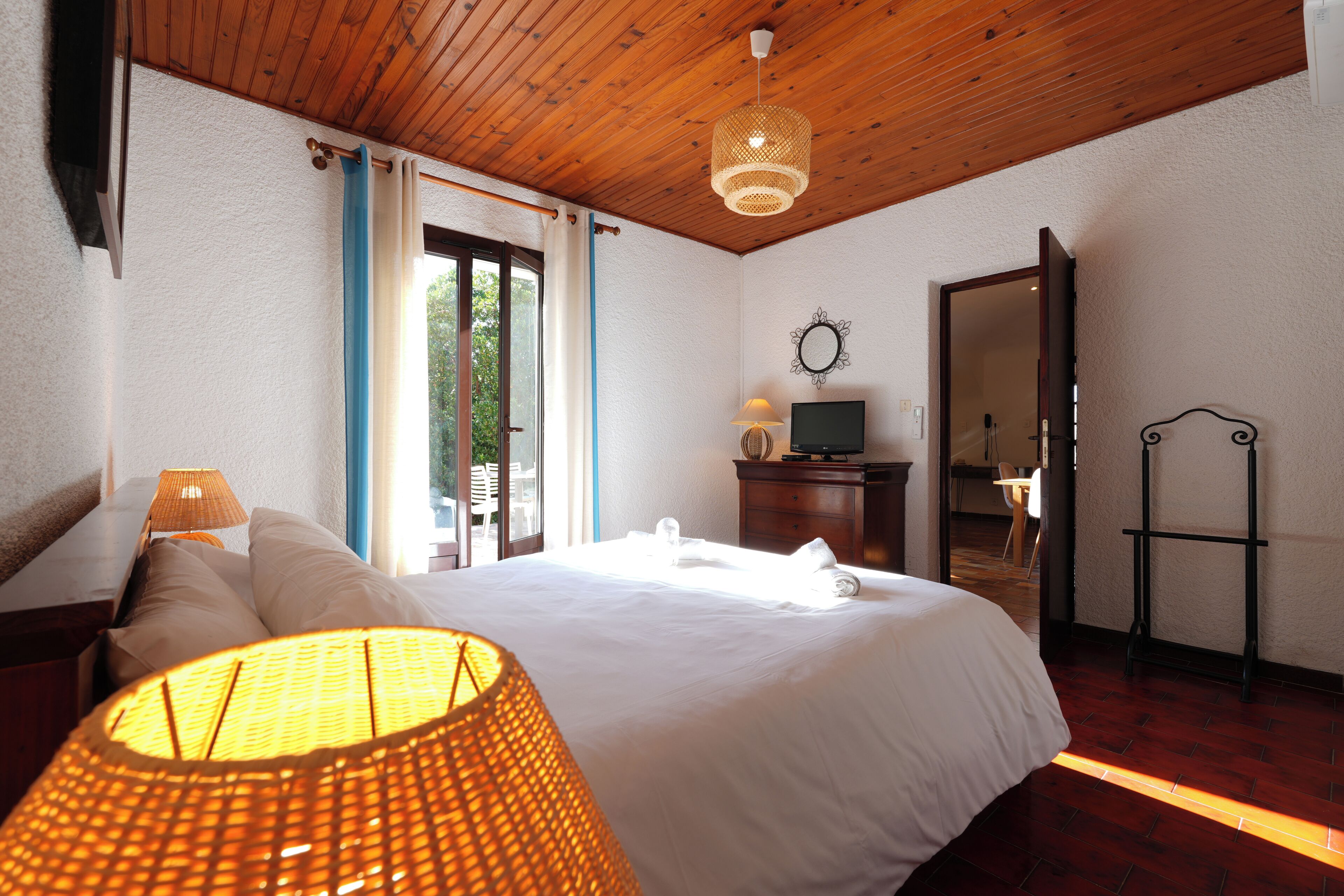A hotel room with a wooden ceiling, a bed with white linens, a lamp, a TV, and a mirror.