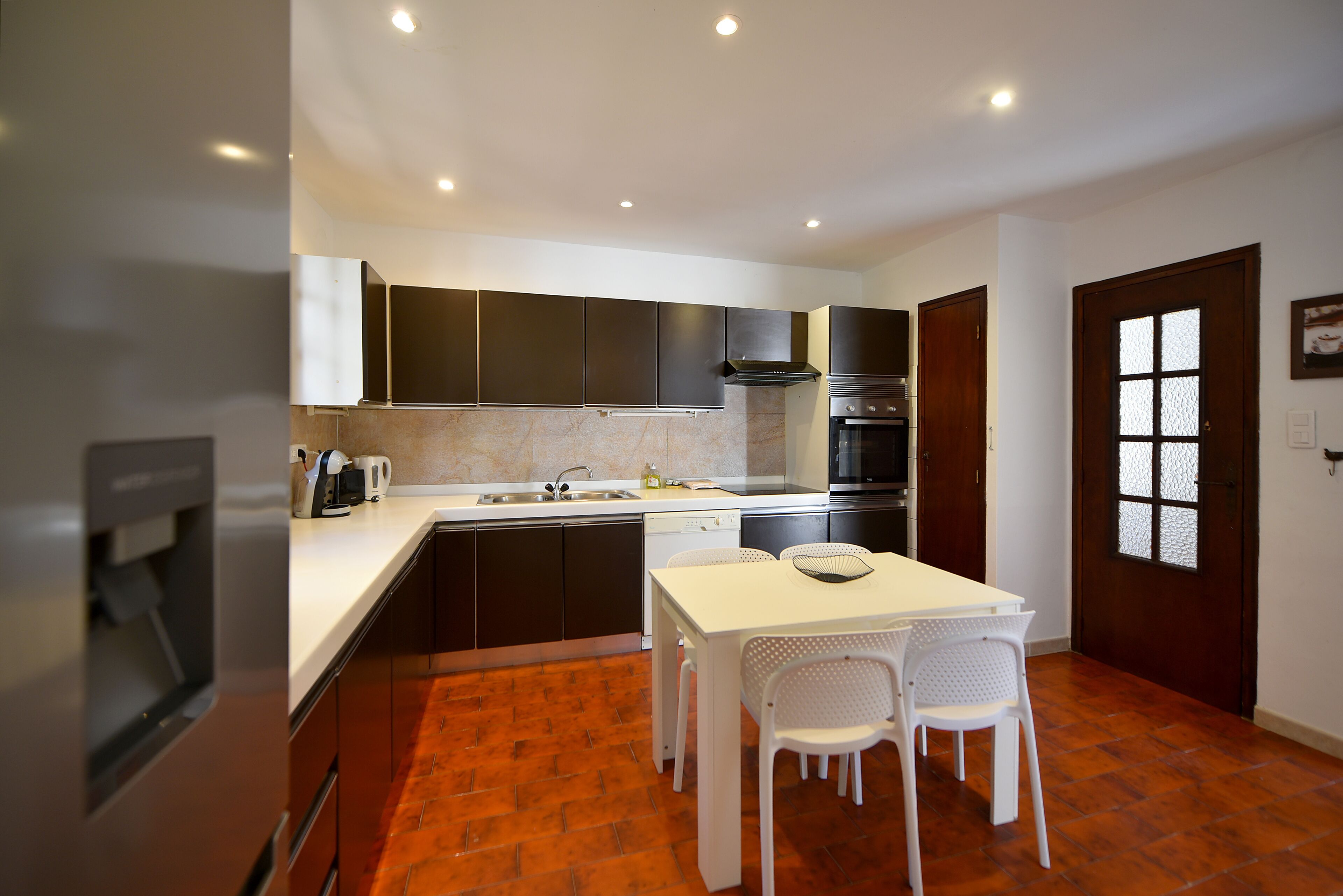 A compact kitchen with dark cabinets, a white countertop, a built-in oven, and a dining area with a white table and chairs.