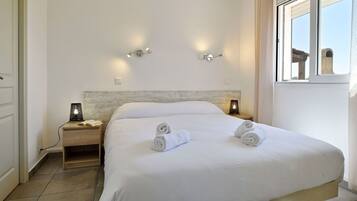 A neatly made bed with two rolled towels, a bedside table with a lamp, and a window with a view of a stone structure.