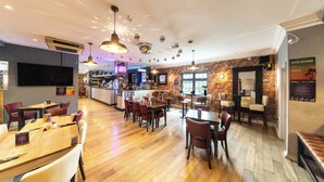 A restaurant with wooden flooring, multiple tables and chairs, hanging pendant lights, a bar area, and brick walls. - The Littleover Lodge Hotel (Derby)
