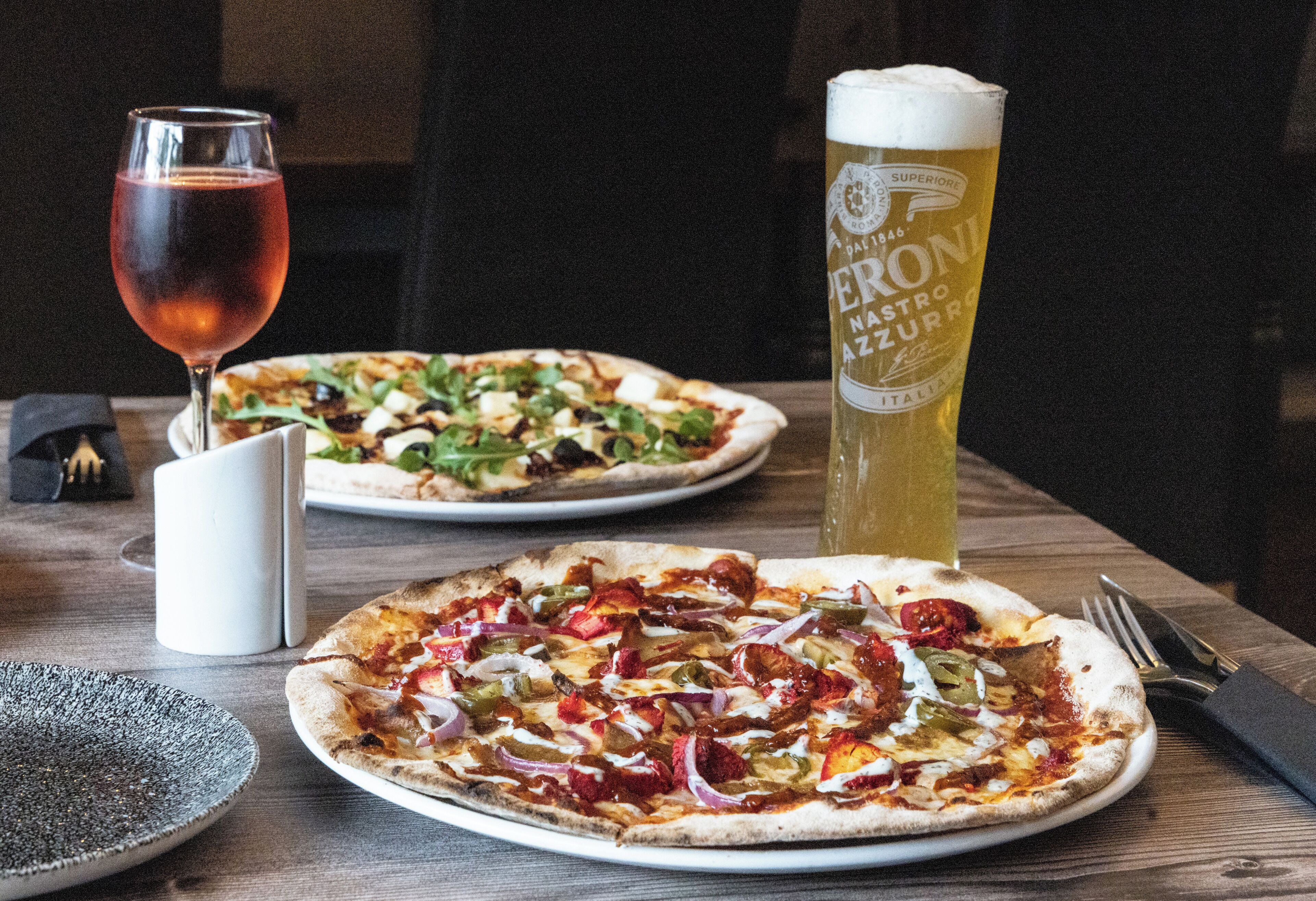 Two pizzas on plates, a glass of beer, and a white mug on a wooden table.