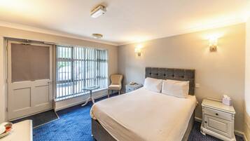 A hotel room with a large bed, a nightstand, a chair, a desk, and a window with blinds.