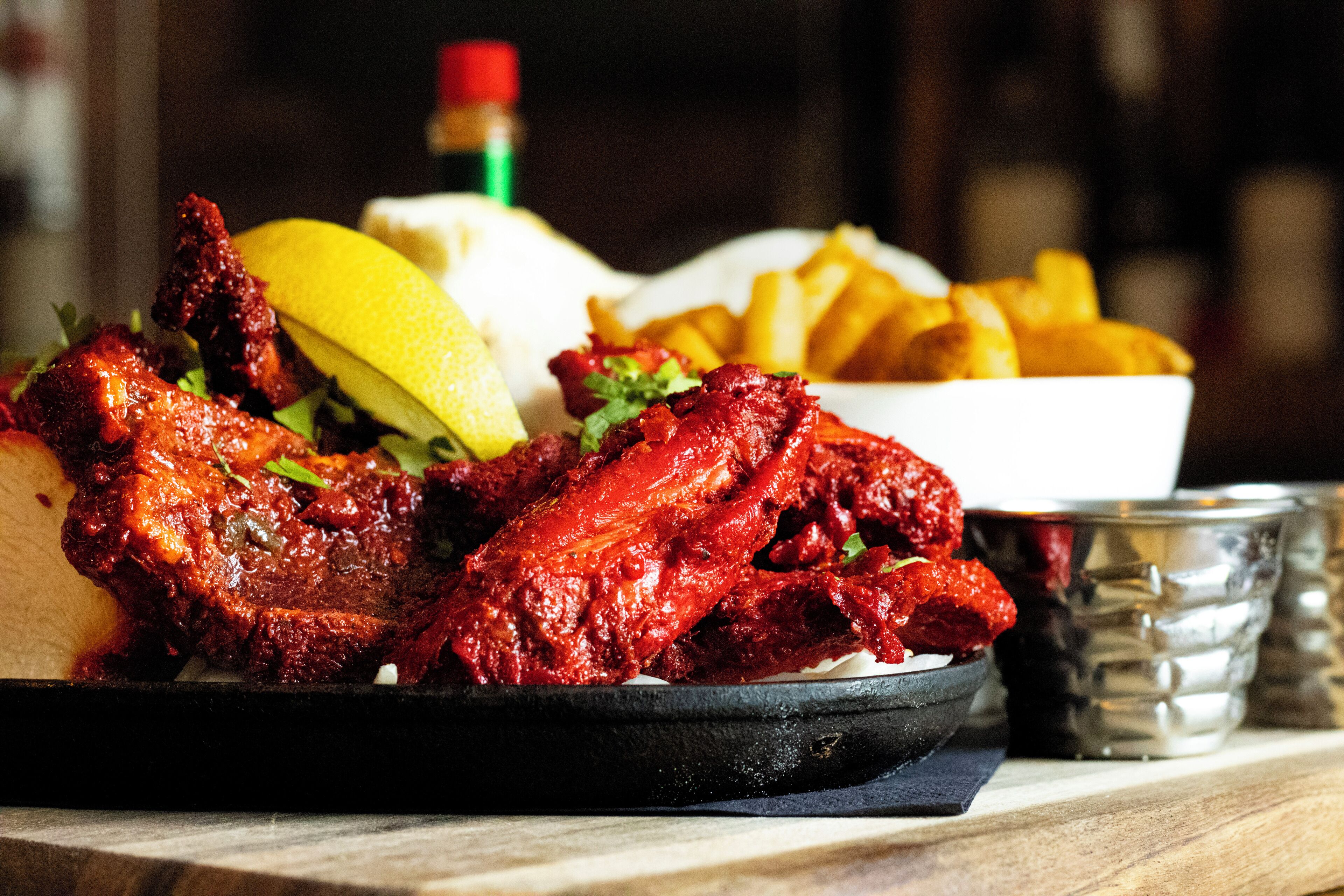 Une assiette d’ailes de poulet rouges et laquées, accompagnées de quartiers de citron et d’une portion de frites dorées.