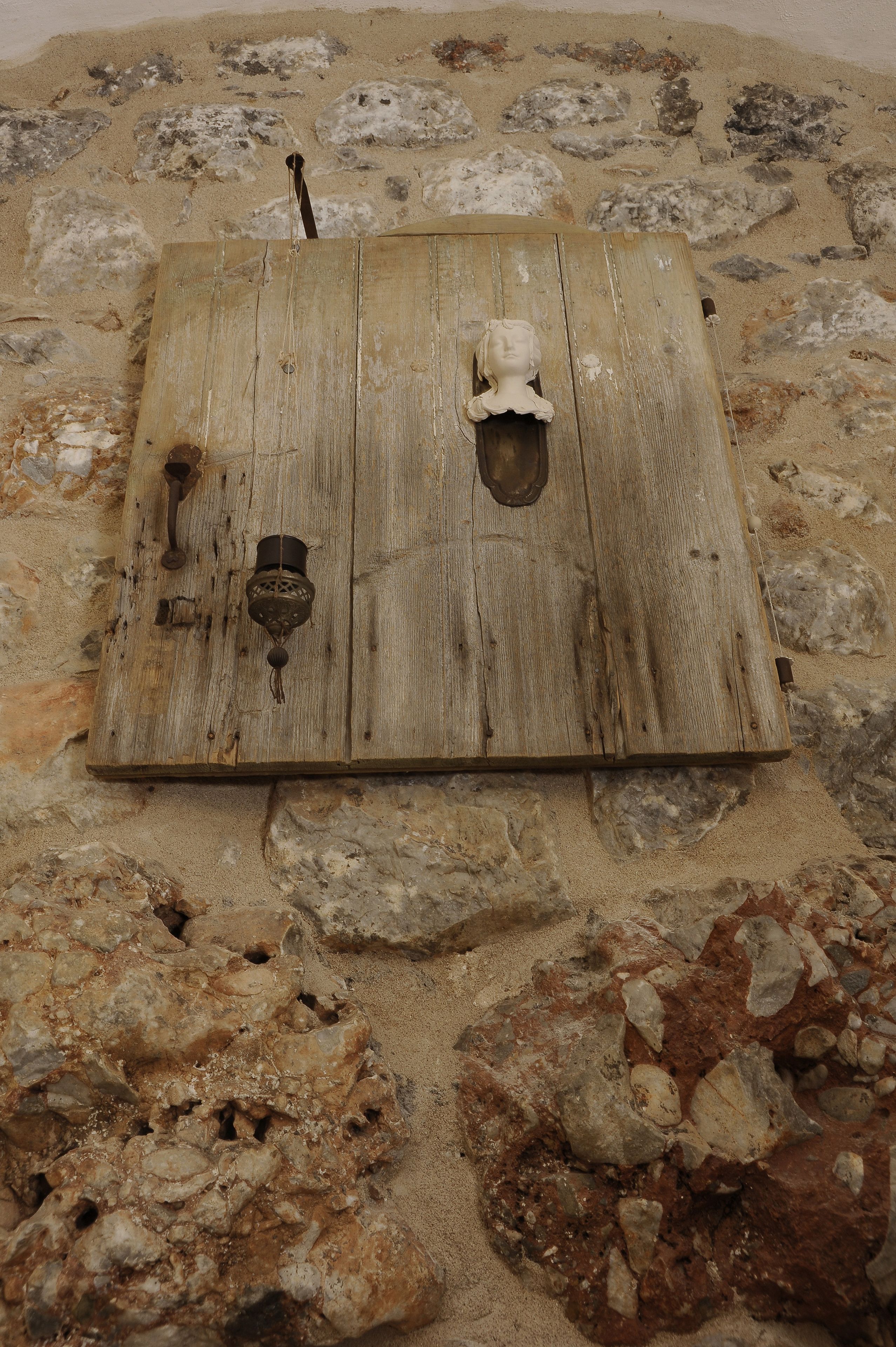 Una puerta de madera rústica con un picaporte de metal y un pequeño objeto metálico adherido, sobre un muro de piedra.