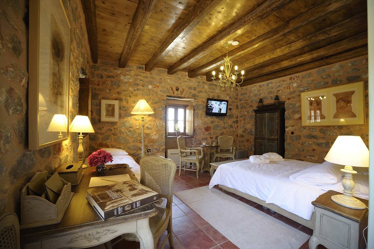 A rustic room with stone walls, wooden beams, a bed, a desk with a lamp, a television, and a chair.