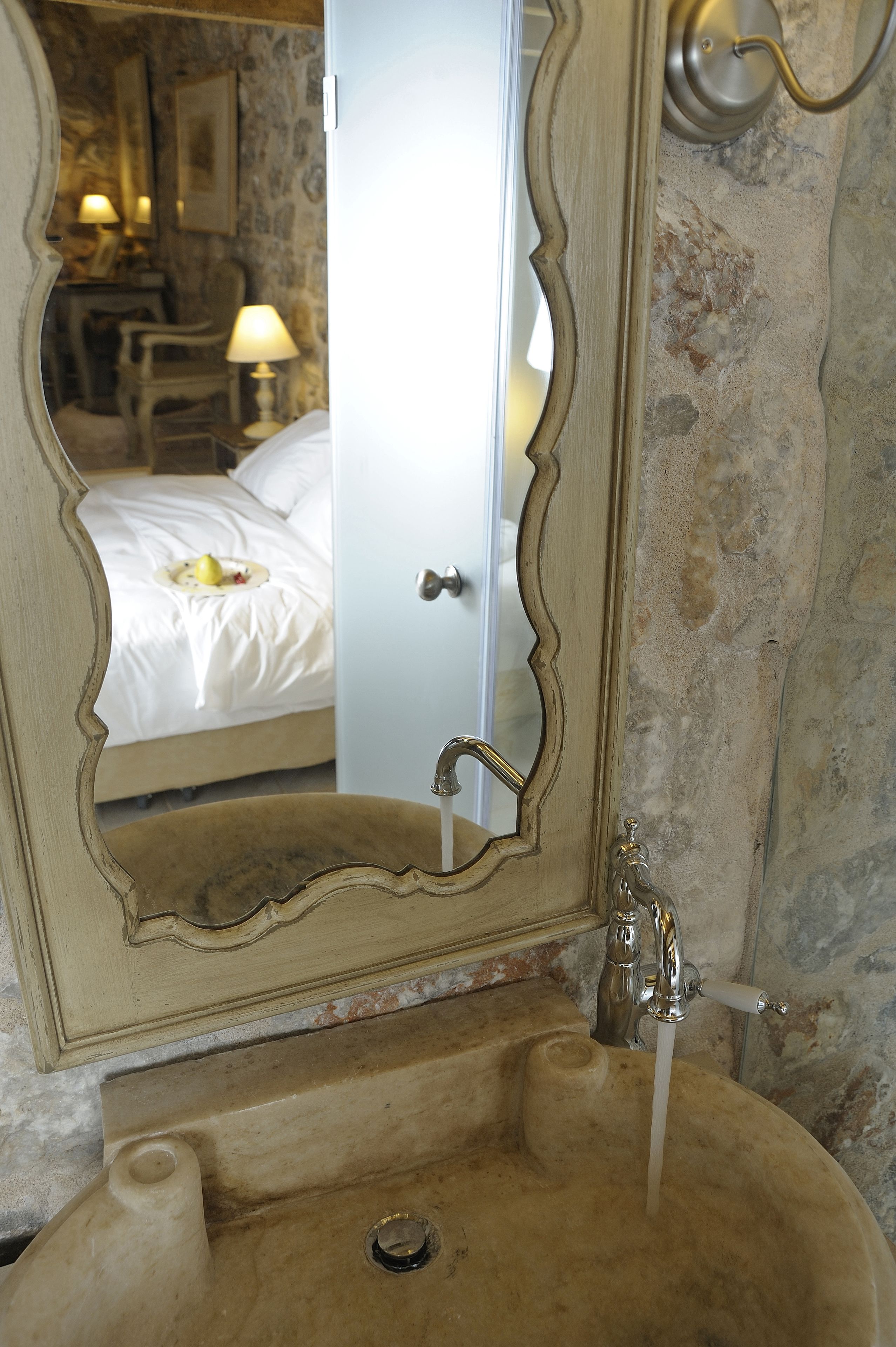 A bathroom with a marble sink, a mirror reflecting a bed and a lamp, and a door with a handle.