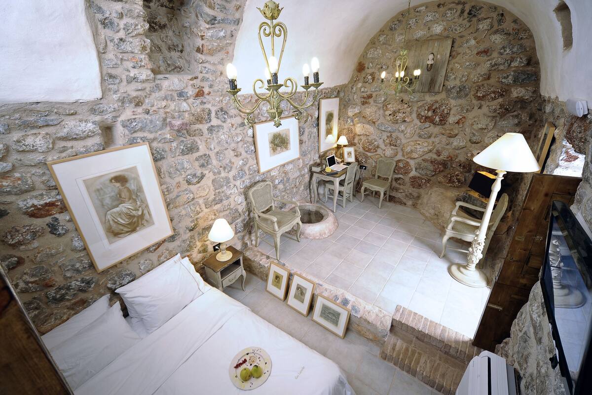 A stone-walled room with a bed, a desk with a lamp, and framed pictures on the walls.