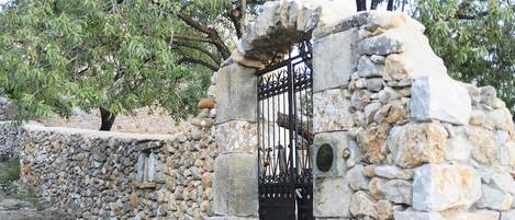 Un portón de piedra con una puerta metálica, rodeado de una pared de piedra y árboles.